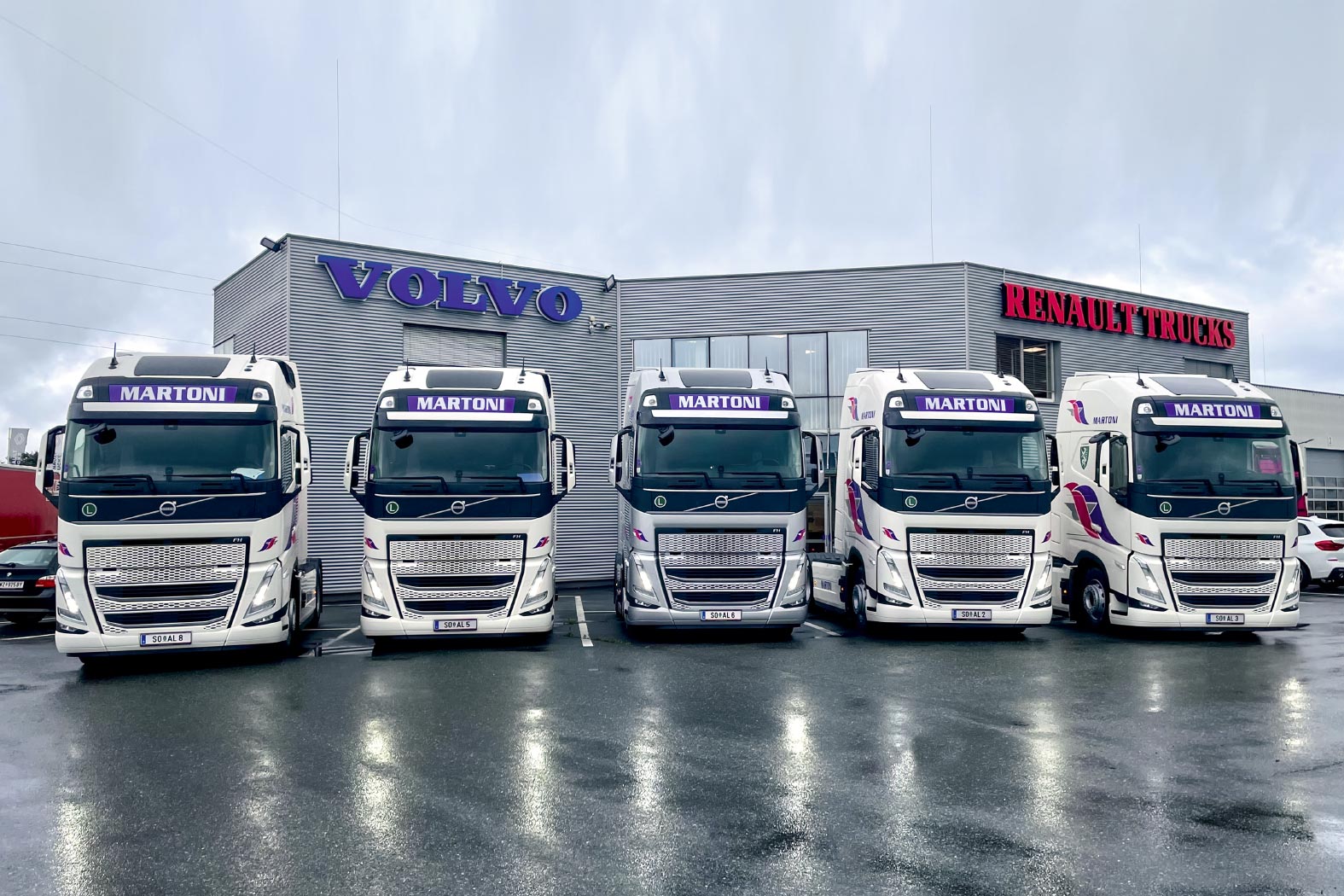 F&uuml;nf wei&szlig;e Volvo-LKWs mit &sbquo;Martoni&lsquo;-Beschriftung vor einem Geb&auml;ude mit Volvo und Renault Trucks Logos.