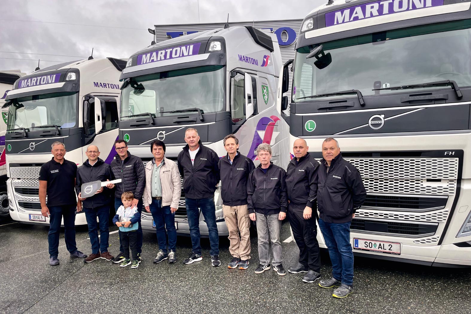 Gruppe vor Volvo-LKWs mit Martoni-Schriftzug, eine Person h&auml;lt symbolischen Schl&uuml;ssel, sowie Kind dabei.