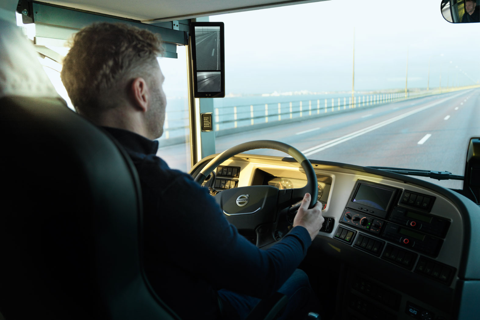 Fahrer in einem Volvo-LKW &uuml;berquert eine lange Br&uuml;cke bei Tageslicht, mit digitalem R&uuml;ckspiegel.