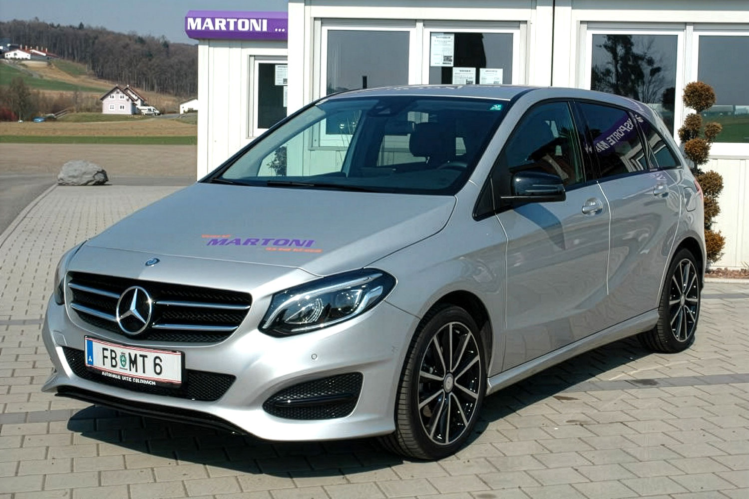Ein silberner Mercedes-Benz-Kompaktwagen auf einem Parkplatz vor einem Geb&auml;ude mit der Aufschrift 'MARTONI'.
