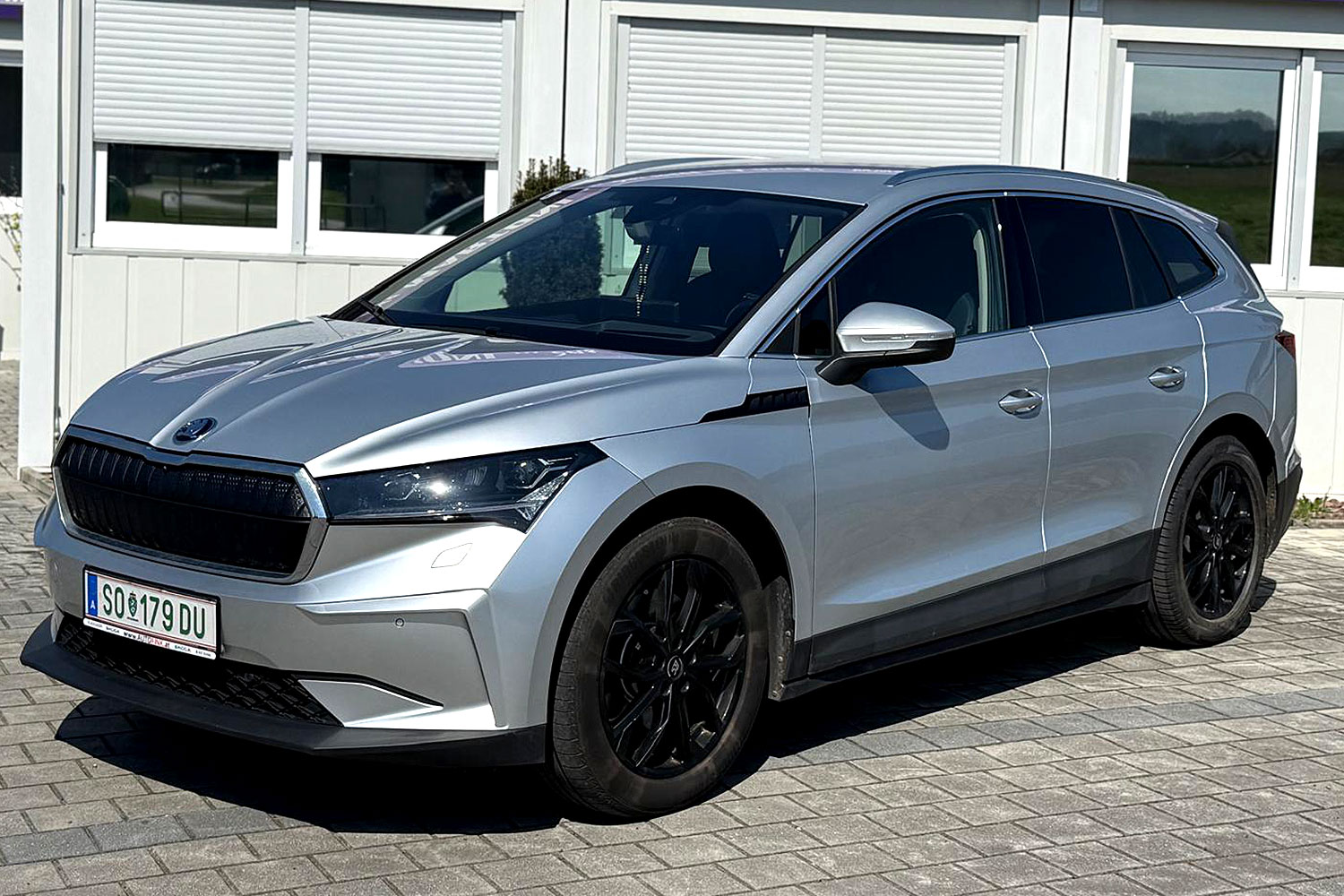 Silberner Skoda Enyaq SUV auf Parkplatz vor Geb&auml;ude mit geschlossenen Jalousien.