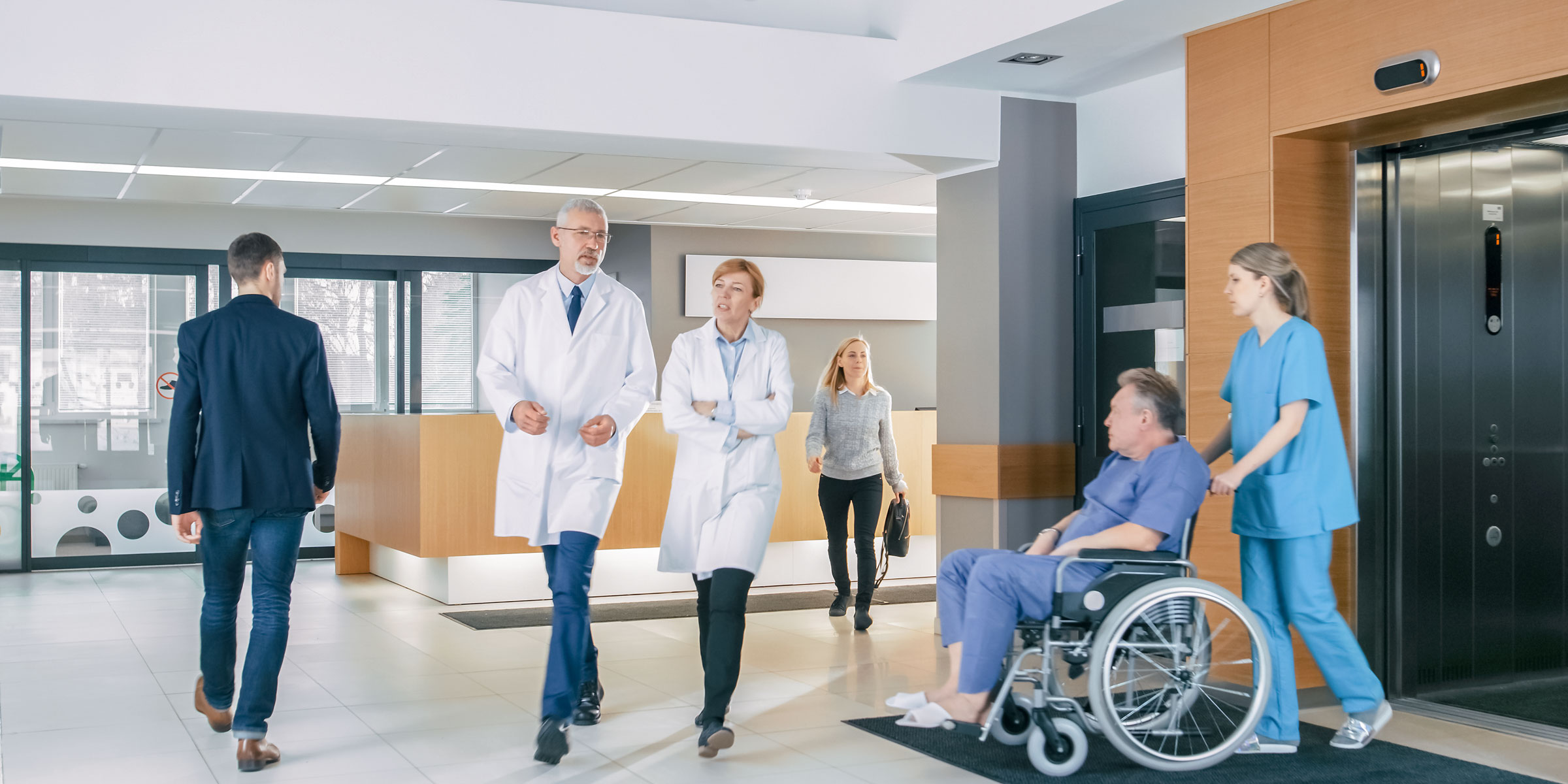 Mehrere Personen in Krankenhauslobby, darunter &Auml;rzte und eine Krankenschwester mit Patient im Rollstuhl.